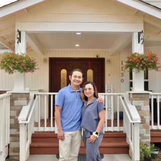 A smiling couple standing in front of a house on the porch.