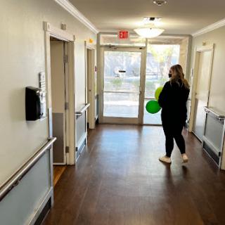 A woman holding a green balloon stands in a well-lit hallway with doors on either side.