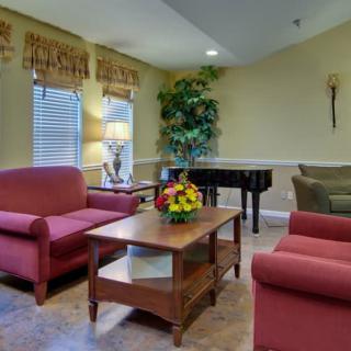 A cozy living room featuring a red armchair and sofa, with a wooden coffee table adorned with a colorful flower arrangement. A piano is placed in the corner, complementing the warm decor.