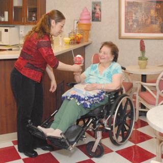 A caregiver assists an elderly woman in a wheelchair, bringing her a treat in a cozy indoor setting.