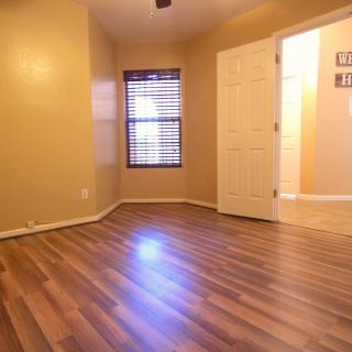 A warm-toned empty room with hardwood flooring, featuring window blinds and an open doorway.