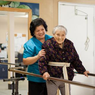 A caregiver assists an elderly person during a physical therapy session.
