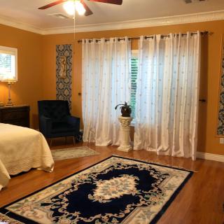 A cozy bedroom with warm tones, featuring a bed, armchair, decorative rug, sheer curtains, and a potted plant.