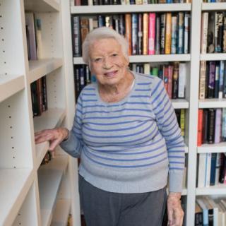 An elderly woman smiling while standing in front of a bookshelf.