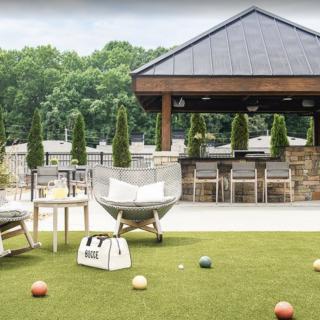 A serene outdoor patio area featuring lounge chairs, a bar space, and bocce balls on a grassy surface, surrounded by lush greenery.