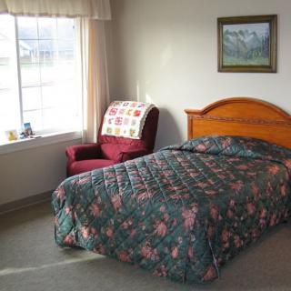 A cozy bedroom featuring a single bed with floral bedding, an armchair by the window, and a landscape painting on the wall.