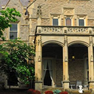 A historic stone mansion with arched windows and a porch featuring columns, surrounded by a garden.