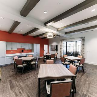 A modern dining area with several tables and chairs, featuring wood flooring, an orange accent wall, ceiling beams, and a kitchenette.