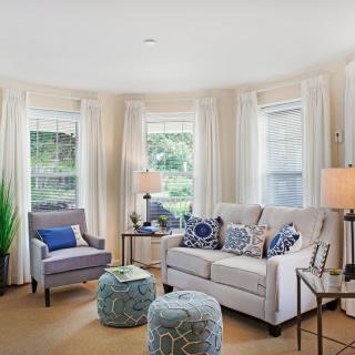 A cozy living room with a sofa and armchair, adorned with decorative pillows and surrounded by windows with sheer curtains.