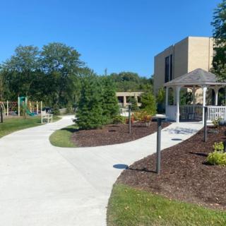A scenic outdoor area with a pathway leading to a gazebo and playground, surrounded by lush greenery and modern buildings.