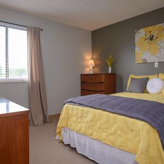 A cozy bedroom with a yellow and gray color scheme, featuring a bed, dresser, lamp, and window with curtains.