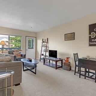 A cozy living room with a sofa, coffee table, TV, desk, and shelves, featuring natural light from a large window.