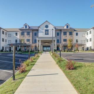 A modern hotel building with a wide entrance pathway, surrounded by a neatly maintained parking area and landscaping on a sunny day.