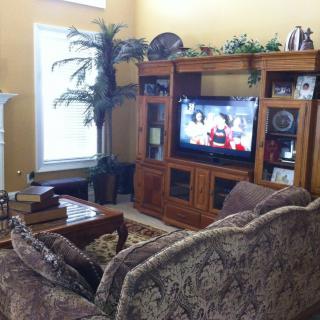 A cozy living room with a large entertainment center and television, featuring a patterned couch and a coffee table set against warm yellow walls.