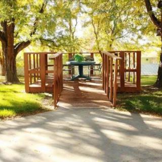 A scenic view of a park with a wooden bridge leading to a picnic area under the shade of trees.