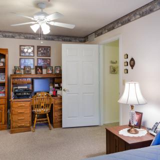 A cozy bedroom with a wooden desk, chair, and computer setup, illuminated by a table lamp. A ceiling fan hangs from the ceiling.