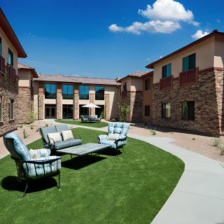 A sunny courtyard with outdoor seating, featuring a sofa and chairs on a grassy area surrounded by buildings.