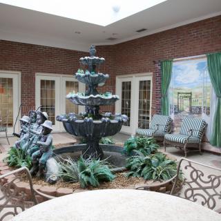 A serene indoor courtyard featuring a multi-tiered fountain surrounded by statues, plants, and comfortable seating in a well-lit space with brick walls.