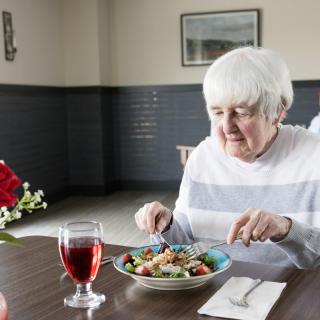 An elderly woman enjoys a meal at a table set with a salad and a glass of red drink.