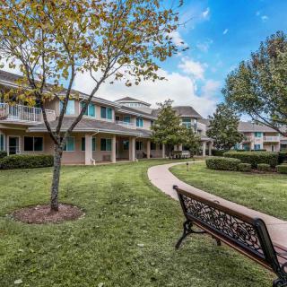 A peaceful courtyard with apartment buildings, featuring a grassy area, trees, a pathway, and a bench under a cloudy sky.