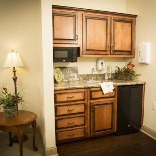 A small kitchenette with wooden cabinets, a countertop, a sink, and a microwave. A lamp and side table are placed nearby.