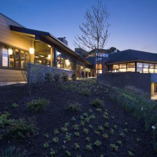 A modern building with illuminated exteriors at dusk, featuring landscaped surroundings.