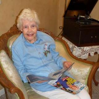 An elderly woman smiles while sitting in a vintage armchair, holding a magazine in a cozy living room.