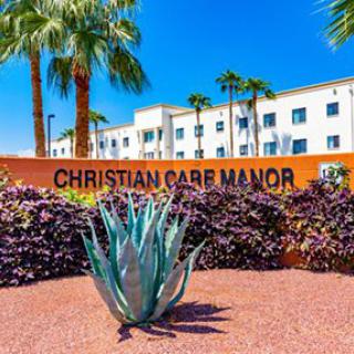 The entrance to Christian Care Manor with palm trees and a succulent in the foreground under a clear blue sky.