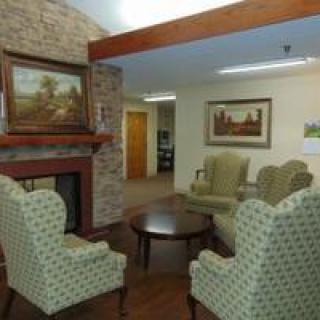 A cozy living room with armchairs arranged around a small table, featuring a brick fireplace and framed artwork on the walls.