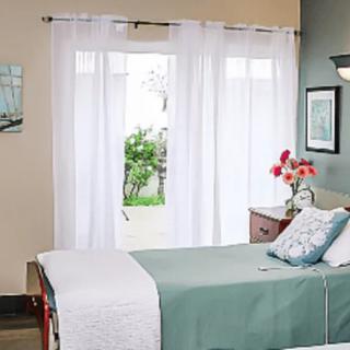 A cozy bedroom with a neatly made bed, light curtains, and a view of greenery outside.