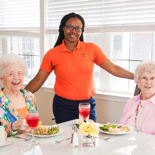 A caregiver posing with two elderly women enjoying a meal together.