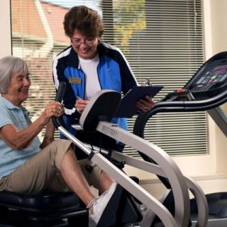 An elderly woman exercises on a machine with assistance from a trainer.