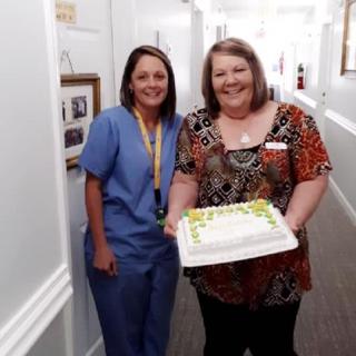 Two women in a hallway, one holding a cake, smiling for the photo.