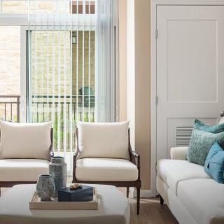 A cozy living room with a white sofa, two chairs, and a coffee table decorated with vases, set beside large windows with blinds.