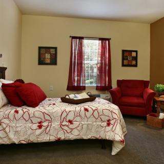 A cozy bedroom with a bed featuring floral bedding, a matching red chair, and decorative curtains.
