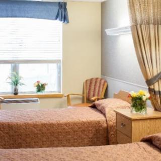A hospital room with two neatly made beds, separated by a curtain, featuring a window with blinds and bedside tables.