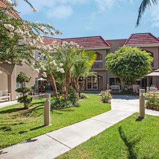 A beautifully landscaped courtyard with a central pathway surrounded by lush greenery and trees, adjacent to a building under a clear blue sky.