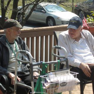 Two elderly men sitting on a porch, engaged in conversation and enjoying the outdoors.