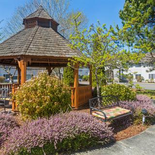 A charming garden with a wooden gazebo surrounded by vibrant shrubs and flowers, featuring a bench and a paved path leading through the landscaped area.