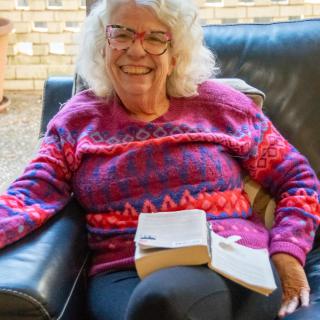 A woman with glasses and a colorful sweater sits on a couch with an open book, smiling at the camera.