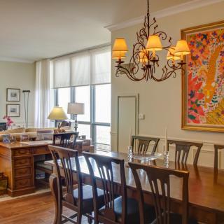 A spacious dining room with a wooden table and chairs, illuminated by an ornate chandelier. A colorful painting decorates the wall.