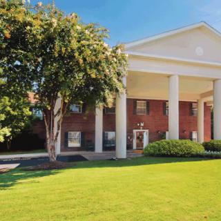 A brick building with white columns on a sunny day, surrounded by a green lawn and trees.