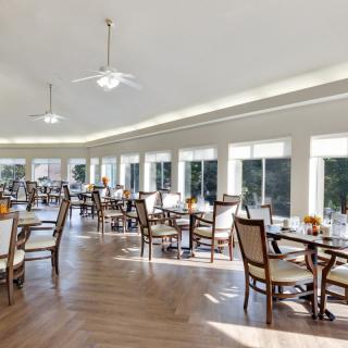 A spacious and bright dining room with multiple tables and chairs, featuring large windows and ceiling fans.