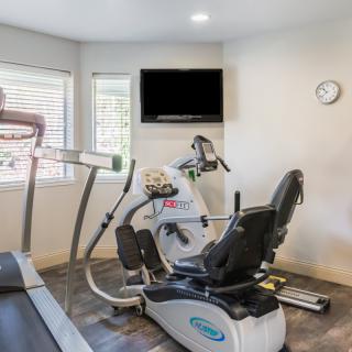 A small fitness room equipped with a treadmill and a recumbent bike, featuring a wall-mounted television and a clock.
