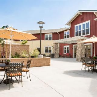 A sunny outdoor patio area with tables, chairs, and umbrellas in front of a red building.