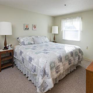 A cozy bedroom featuring a queen-sized bed with decorative bedding and bedside lamps, illuminated by natural light.