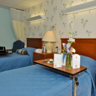 A hospital room with two neatly made beds, featuring floral wallpaper and a small table with flowers and cards.