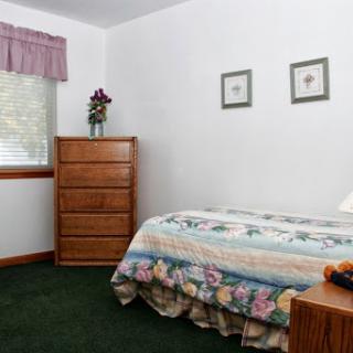 A cozy and simple bedroom featuring a floral bedspread, wooden dresser, and green carpeting.