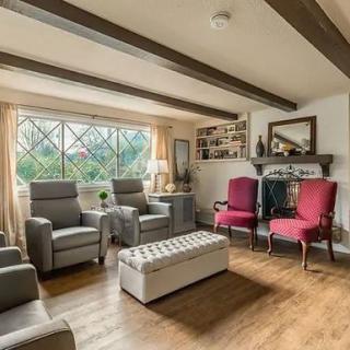 A cozy living room with comfortable armchairs, a wooden floor, and a beamed ceiling, illuminated by natural light from large windows.