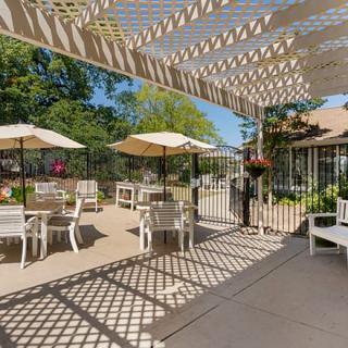 A sunny outdoor patio area with tables, chairs, and umbrellas under a pergola.
